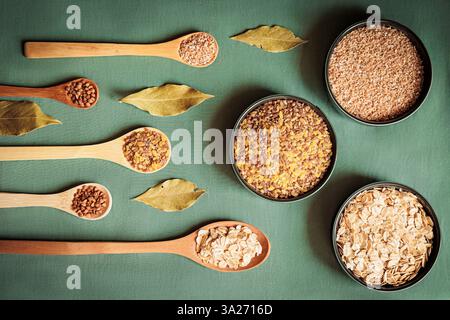 Giacenza piatta di cereali assortiti e cereali in cucchiai e ciotole di legno, decorata con foglie di alloro su sfondo verde. Concetto di alimenti rustici e biologici, Foto Stock