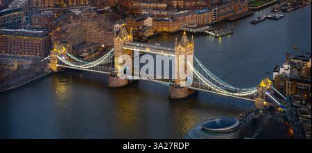 Il Tower Bridge si erge maestosamente sul Tamigi a Londra, illuminando di luci al calare della sera. I battelli navigano sotto mentre lo skyline della città risplende di vita. Foto Stock