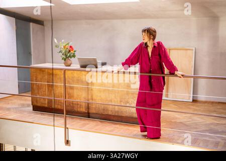 Una donna con un abito rosso si trova accanto a una ringhiera in un loft contemporaneo, guardando in lontananza. Il caldo pavimento in legno, il decor minimalista e la luce naturale soffusa evocano un senso di creatività e indipendenza Foto Stock