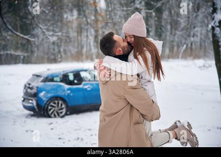 Sorridere, divertirsi. Coppia felice che fa una passeggiata nella foresta invernale. L'auto blu è parcheggiata. Foto Stock