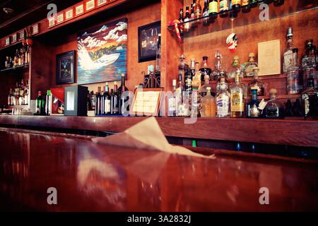 Un bar in legno con una superficie lucida che mostra una varietà di bottiglie di liquori sugli scaffali. Un colorato dipinto di una barca è esposto sulla parete, addin Foto Stock