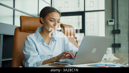 giovane donna sorridente che lavora su un laptop a una scrivania in un ufficio luminoso e moderno con documenti, grafici e smartphone, circondato da mobili eleganti Foto Stock