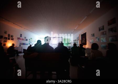 Una sala poco illuminata con persone sedute che guardano una presentazione su un grande schermo. Le pareti sono adornate da varie fotografie, creando un'a artistica Foto Stock