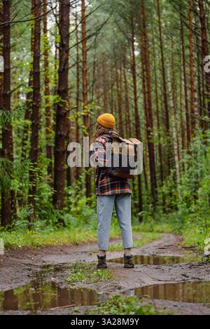 Una giovane escursionista che indossa uno zaino e un berretto giallo si sta godendo una tranquilla passeggiata su un sentiero fangoso della foresta dopo la pioggia, circondato da lussureggianti alberi verdi Foto Stock