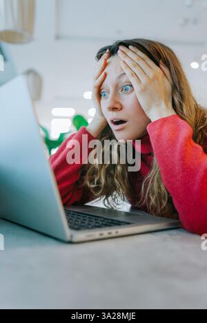 Donna stressata con la testa in mano, che fissa lo schermo del notebook mentre riceve cattive notizie su un attacco informatico o un errore di sistema Foto Stock