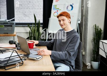 Un giovane uomo con i capelli rossi si impegna attivamente con il suo laptop in un ambiente moderno. Foto Stock