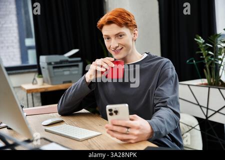 L'uomo dai capelli rossi sorride mentre sorseggia una tazza di caffè mentre lavora sui suoi compiti. Foto Stock