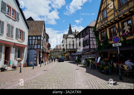 Kaysersberg, Francia - 15 giugno 2024: Una pittoresca strada di Kaysersberg caratterizzata da case colorate e in legno e da ristoranti all'aperto. Foto Stock