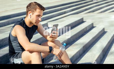L'atleta con cuffie wireless e il tracker per il fitness tiene in mano una bottiglia d'acqua e uno smartphone, si siede sulle scale Foto Stock