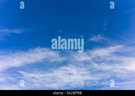 Un cielo sereno mette in mostra morbide nuvole di velluto su uno sfondo blu brillante. Una piccola luna crescente può essere vista, aggiungendo un tocco di tranquillità al giorno Foto Stock