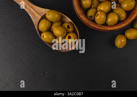 Le olive verdi si trovano in una ciotola di legno accanto a un cucchiaio di legno, mostrando il loro colore vivace su uno sfondo scuro. Questa impostazione sottolinea la natura Foto Stock