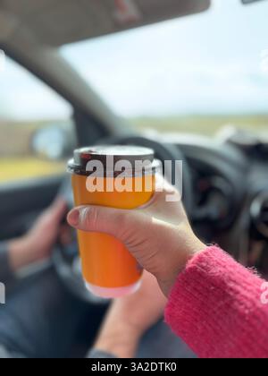 Una donna che regge una tazza di caffè arancione contro il volante di un'auto, simboleggiando viaggi, takeout e avventure. Donna che tiene la tazza di caffè in auto. Foto di alta qualità Foto Stock