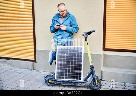 La persona di sesso maschile contiene un pannello solare fotovoltaico, per ricaricare uno scooter elettrico, utilizzando lo smartphone. Integrazione dell'energia solare come fonte di energia sostenibile, promuovendo un trasporto urbano ecocompatibile. Foto Stock