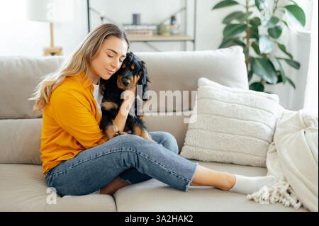 Felice e affascinante donna caucasica in abiti casual, abbraccia delicatamente il suo cane nero e marrone su un divano a casa, esprimendo amore e calore, un momento emozionante di legame tra un animale domestico e il suo proprietario Foto Stock
