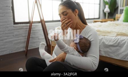 Donna che sbadiglia mentre tiene la bambina in camera da letto accogliente mostrando la madre stanca con amore in casa. Foto Stock
