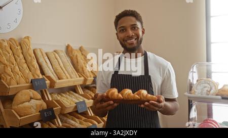 Bel giovane sorridente all'interno di una panetteria che tiene in mano un vassoio di croissant appena sfornati con una varietà di pane sugli scaffali sullo sfondo Foto Stock