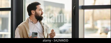 Un bell'anziano con un abbigliamento casual tiene una tazza di caffè e guarda all'esterno con riflessi. Foto Stock