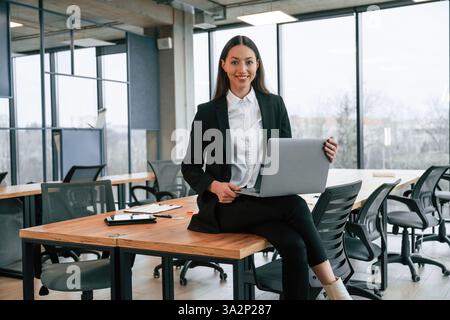 Seduti sul tavolo con il laptop in ginocchio. Una donna d'affari è al chiuso in ufficio. Foto Stock