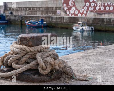 Una corda spessa e resistente agli agenti atmosferici avvolta intorno a un dissuasore arrugginito in un porto, con barche e un badile ricoperto di murales sullo sfondo. Foto Stock