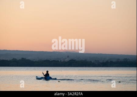 Pagaiando tranquillamente sull'acqua serena, un kayak gode del tramonto tranquillo con colline lontane in vista. Foto Stock