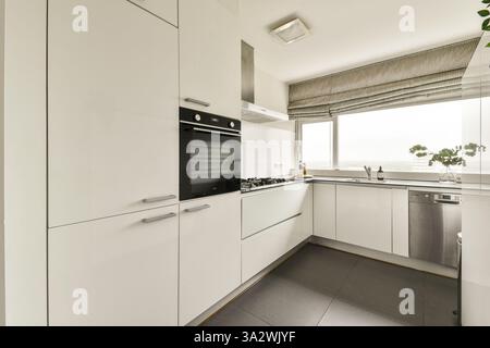 Eleganti mobili bianchi con elettrodomestici moderni, grandi finestre con luce naturale e un'estetica contemporanea definiscono questo elegante spazio cucina. Foto Stock