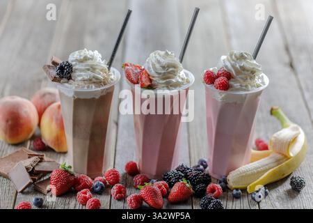 Tre deliziosi frullati con panna montata, frutti di bosco freschi, cioccolato e frutta, serviti in tazze alte su un rustico tavolo di legno con ingredienti. Foto Stock