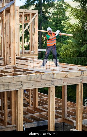 Edificio carpentiere casa con telaio in legno vicino alla foresta. Uomo barbuto con occhiali che reggono una grande tavola sulla spalla, vestito con abiti da lavoro e casco. Concetto di costruzione moderna e sostenibile. Foto Stock
