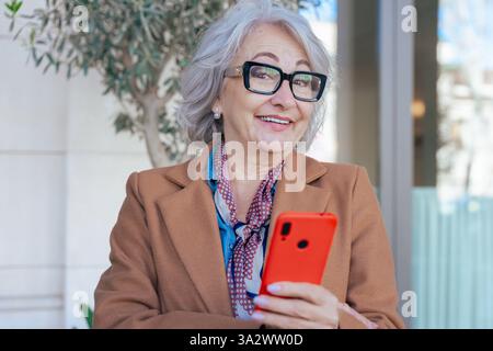 Donna anziana che guarda la fotocamera con un sorriso e tiene in mano uno smartphone rosso all'aperto. Foto Stock