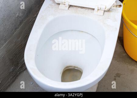 Un gabinetto sporco e poco pulito si trova proprio accanto a un vecchio secchio giallo, che probabilmente viene utilizzato per la pulizia del bagno Foto Stock