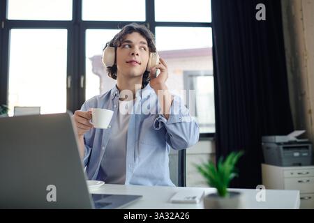 Il giovane uomo si prende un momento per rilassarsi e sorseggiare un caffè mentre lavora in un'elegante area di lavoro. Foto Stock
