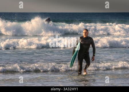 Un surfista maschio maturo che porta la sua tavola da surf e si allontana dal mare dopo una sessione di surf al Fistral di Newquay in Cornovaglia nel Regno Unito in Europa Foto Stock