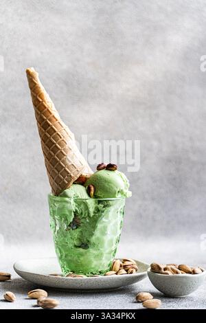 Una rinfrescante tazza di gelato al pistacchio con noci in un bicchiere testurizzato, condito con un cono croccante di waffle e guarnito con pistacchi interi, su una Foto Stock
