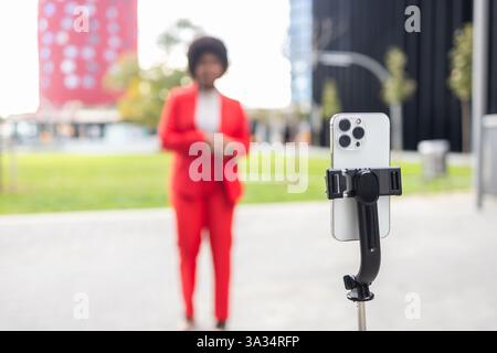 Una donna con una vivace tuta rossa sta all'aperto, concentrandosi sulla registrazione con il suo smartphone su un cavalletto. L'ambiente e' urbano, con edifici moderni in Foto Stock