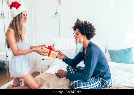 Una scena gioiosa di una coppia che si scambiava un regalo confezionato in una camera da letto accogliente. La donna indossa un cappello di Babbo Natale, che simboleggia l'allegria festiva e lo spirito festivo. Foto Stock