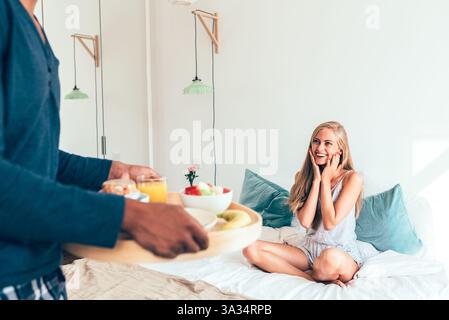 Una donna reagisce con gioia a una colazione a sorpresa a letto in una mattinata di sole. Un uomo porta un vassoio con frutta, succo di frutta e fiori, creando uno sta perfetto Foto Stock