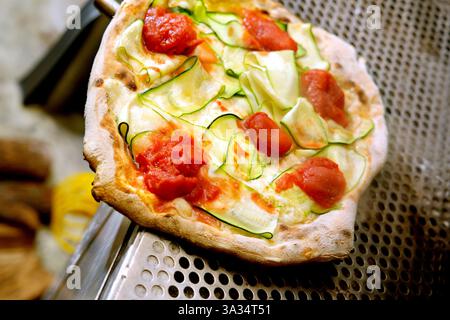 Una pizza vegetariana appena sfornata condita con fette di zucchine, pezzi di pomodoro, sollevati da una spatola da un vassoio per pizza perforato Foto Stock