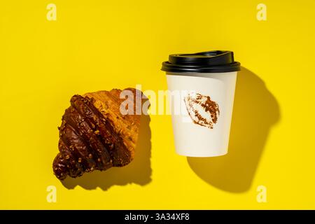 Vista dall'alto di un croissant al cioccolato abbinato a una tazza da caffè da asporto, che presenta una macchia di rossetto, il tutto adagiato su un vivace dorso giallo Foto Stock