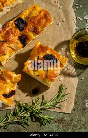 Fette di focaccia appena sfornate con rosmarino e pomodori secchi su carta pergamena, accompagnate da un rametto di rosmarino e da un sma Foto Stock