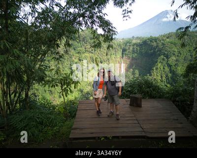 Una coppia allegra in piedi su una piattaforma di legno alla cascata Kaspa Biru, con il maestoso Monte Semeru e la lussureggiante foresta sullo sfondo. Lo sono Foto Stock