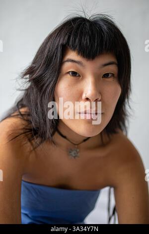 Un ritratto ravvicinato di una giovane donna asiatica con capelli scuri, bangs e piercing facciali. Indossa una collana con girocollo e un top blu senza spalline, che guarda Foto Stock