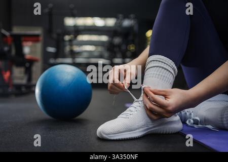 Un primo piano di mani di donna che lega scarpe bianche atletiche su un pavimento di palestra, con una palla blu nelle vicinanze. L'immagine acquisisce la preparazione prima di Foto Stock