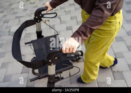 Una donna con paralisi cerebrale utilizza un camminatore per la mobilità come sostegno mentre cammina su un sentiero lastricato. L'immagine mostra un primo piano delle mani che afferrano le maniglie Foto Stock