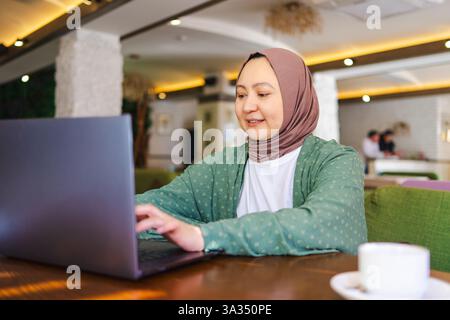 Una donna musulmana che indossa un hijab, seduta in un accogliente bar, che utilizza un laptop. L'ambiente e' moderno e invitante, riflettendo una miscela di tradizionale e con Foto Stock