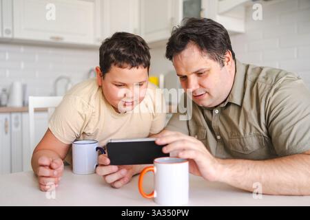Padre e figlio si siedono al tavolo della cucina, entrambi concentrati su uno smartphone. Condividono tempo di qualità, sorseggiando tazze e godendosi un momento di digital ent Foto Stock