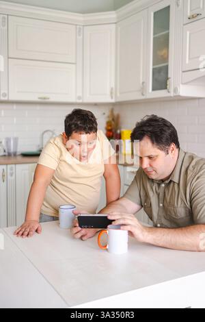 Padre e figlio sono fidanzati con uno smartphone a un tavolo da cucina. Il figlio sembra sorpreso mentre guardano qualcosa insieme, godendosi il loro tempo indoo Foto Stock