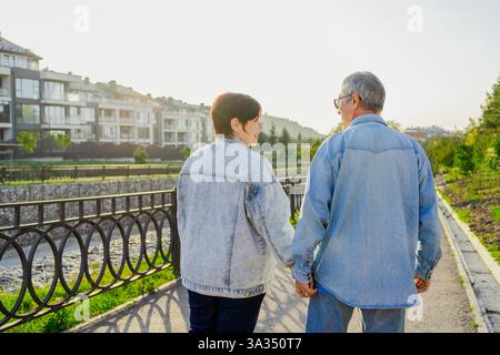 Una coppia di anziani cammina mano nella mano lungo un sentiero illuminato dal sole, indossando giacche in denim. Godono di un momento tranquillo vicino a un fiume, con appartamenti moderni Foto Stock