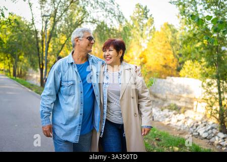 Una coppia di anziani sorridenti gode di una piacevole passeggiata in un parco illuminato dal sole. Camminano braccio nel braccio, circondati da lussureggianti alberi verdi e da un'atmosfera serena, emb Foto Stock