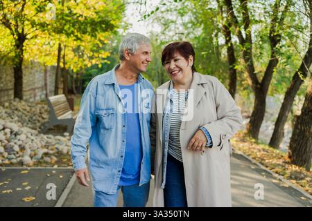 Una felice coppia senior gode di una piacevole passeggiata autunnale in un parco illuminato dal sole. Sorridono e condividono un momento di felicità, circondati da alberi e na Foto Stock