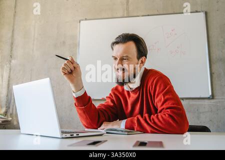 Un uomo con un maglione rosso sorride mentre tiene in mano una penna e si impegna con il suo laptop in un ambiente informale, con una lavagna sullo sfondo. Perfec Foto Stock