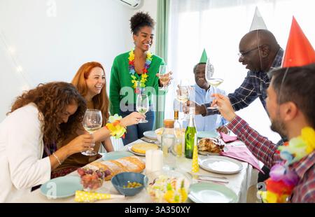 Un gruppo di amici e familiari festeggia con gioia un compleanno a casa, indossando cappelli da festa e colorati leis, brindando con vino e godendosi un bellissimo fu Foto Stock